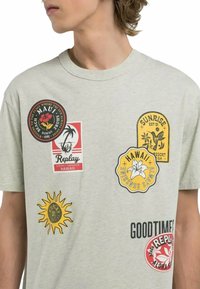 T-shirt in cotone grigio chiaro con diverse toppe colorate a tema hawaiano, tra cui sole, palme e motivi floreali.