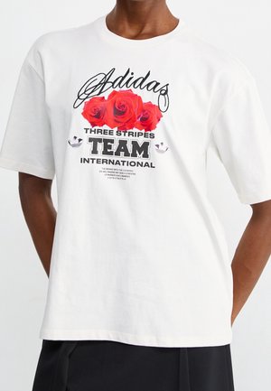 Mand iført hvid Adidas T-shirt med røde roser og teksten "Three Stripes Team International" på fronten.