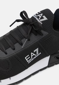Svarta sportiga sneakers med en texturerad stickad ovandel, vit sula och framträdande EA7-logotyp på sidan. Utrustad med platta snören och förstärkt tå.