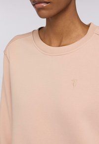 Sudadera de cuello redondo en suave tela color blush. Presenta un cuello acanalado y un pequeño logo bordado en el pecho de color claro. Textura suave.