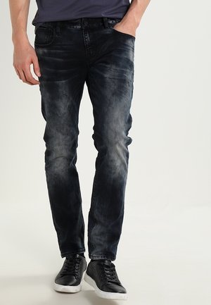 Homme portant un jean noir délavé slim fit et des baskets noires avec des semelles blanches, debout avec une main dans la poche.