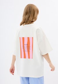 Witte oversized t-shirt met een rechthoekige grafiek in roze en oranje strepen en blauwe tekst op de achterkant. Korte mouwen en een relaxte pasvorm.