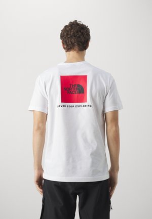 Mann mit lockigem Haar, der ein weißes T-Shirt mit einem roten Quadrat und dem schwarzen "The North Face"-Logo sowie dem Schriftzug "Never Stop Exploring" auf der Rückseite trägt.