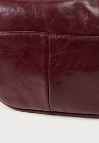 Τσάντα από δέρμα μπορντό με ομαλή υφή, ορθογώνιο σχήμα και ορατές ραφές κατά μήκος των άκρων.