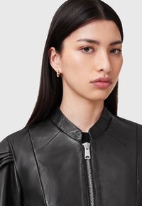 Giubbotto in pelle nera con colletto rialzato, caratterizzato da una zip argentata e da sottili arricciature sulle spalle. Tessuto liscio, elementi di design moderno.