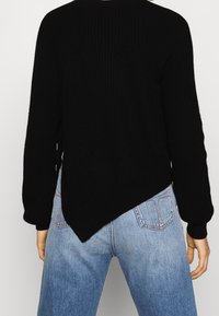 Person klädd i en svart asymmetrisk stickad tröja och blå denimjeans, sedd bakifrån mot en neutral vit bakgrund.