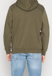Olivgrüner Hoodie mit klassischem Design, ausgestattet mit einer Kapuze und RIP-Bündchen. Der Stoff ist weich und hat eine glatte Textur. Trägt man über hellblauen Jeans.