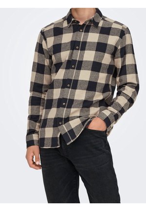 Mann trägt beige und schwarz kariertes Hemd mit langen Ärmeln und schwarze Jeans, eine Hand in der Tasche, vor einfarbigem Hintergrund.