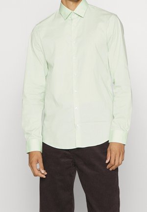 Camicia a maniche lunghe verde chiaro in tessuto morbido, con colletto a punta e chiusura con bottoni, abbinata a pantaloni in velluto a coste marrone scuro.