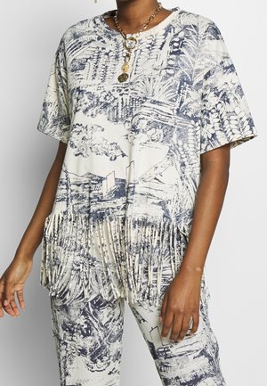 Osoba nosząca białą i granatową, wzorzystą bluzkę z krótkim rękawem z frędzlami na dole oraz pasujące spodnie, uzupełnione złotym łańcuszkiem.