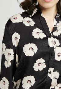 Blouse noire à motifs floraux avec de grandes fleurs blanches rehaussées de détails roses, un col classique et un tissu lisse et texturé avec fermeture à boutons.