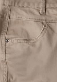 Beige puuvillahousut etutaskulla, näkyvillä saumoilla ja koristeellisella metallikoristeella taskussa. Sujuva pinta.