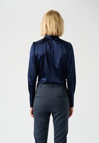 Blusa di raso blu con spalle leggermente abbellite, dettaglio a chiave sulla schiena, abbinata a pantaloni scuri a righe sottili con tasche posteriori.