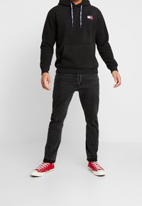 Felpa nera con tasca a marsupio frontale e logo, abbinata a jeans scuri e sneakers rosse. La felpa presenta lacci e polsini a coste.