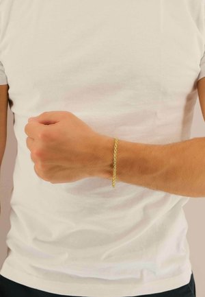 Bracciale a catena in oro indossato al polso, caratterizzato da un design delicato e intrecciato con una finitura liscia, in contrasto con una camicia bianca semplice.