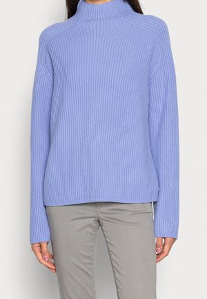 Femme portant un pull à col roulé côtelé violet clair et un pantalon beige, debout devant un fond clair uni.