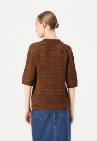 Brun strikket cardigan med korte ermer, intrikate åpne mønstre, rund krage og løs passform. Båret over et denimskjørt.