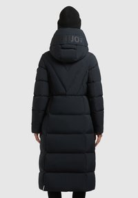Dolga črna puhovka s kapuco, ki ima podložene predele in velik logo na hrbtu. Nosi se čez črne legice in s kapico.