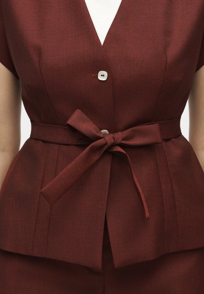 Κοντινή λήψη ενός καλοραμμένου κοντομάνικου σακακιού σε σκουροκόκκινο χρώμα σκουριάς με λευκό κουμπί και ταιριαστή υφασμάτινη ζώνη δεμένη στη μέση.