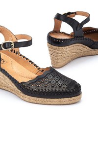 Sandalias negras de cuña con un diseño floral recortado, parte superior de cuero y suela envuelta en yute. Cuentan con una correa al tobillo y detalle de hebilla circular.