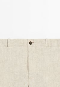 Närbild på beige rutig byxa med knappstängning och bältesöglor på enfärgad vit bakgrund.