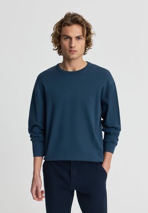 Giovane uomo con capelli ricci, indossa un maglione blu navy a maniche lunghe e pantaloni blu navy, in piedi con una mano in tasca su uno sfondo semplice.