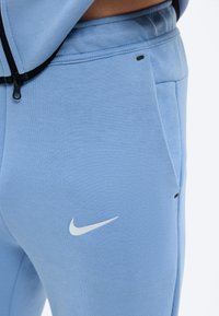 Άτομο που φοράει ανοιχτό μπλε φόρμα Nike με λευκό λογότυπο Swoosh, χέρι στην τσέπη, και μέρος ανοιχτό μπλε μπουφάν με μαύρο φερμουάρ εμφανές.