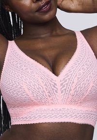 Vaaleanpunainen pitsinen bralette, jossa on V-mallinen kaula-aukko. Siinä on monimutkaisia kuvioita, teksturoitua kangasta ja leveät, pehmeät hihnat. Ei muotoilevaa rautaa.