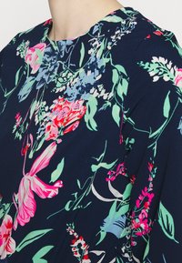 Blusa com padrão floral, apresentando flores vibrantes em tons de rosa, azul e verde sobre um fundo azul-marinho escuro. Decote arredondado e textura de tecido suave.