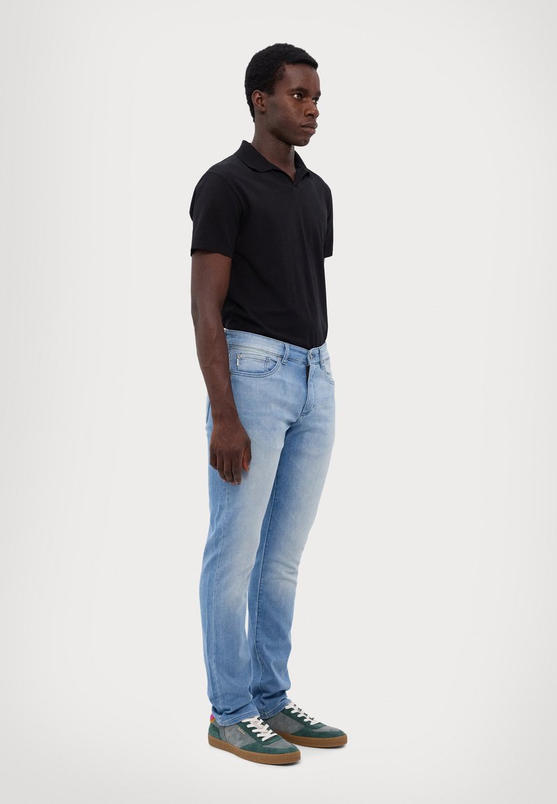 Polo shirt noir à manches courtes, jeans bleu clair et baskets vertes avec lacets blancs. Le modèle se tient de profil contre un fond neutre.