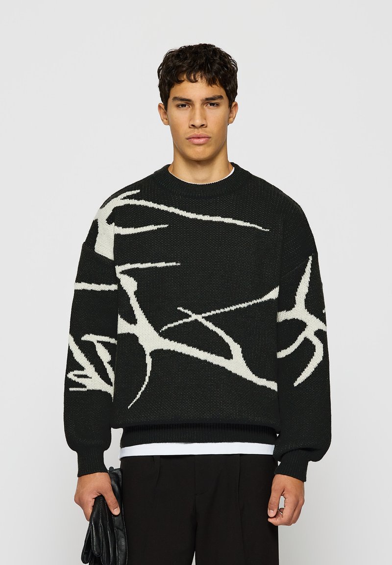 Joven vestido con un suéter negro con patrones abstractos en blanco, pantalones negros, sosteniendo guantes de cuero negros frente a un fondo gris claro liso.