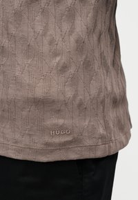 Κοντινή λήψη υφάσματος πλεκτού σε ταουπ απόχρωση με γεωμετρικό σχέδιο και μικρό κεντητό λογότυπο "HUGO" κοντά στο κάτω μέρος πάνω από μαύρο παντελόνι.