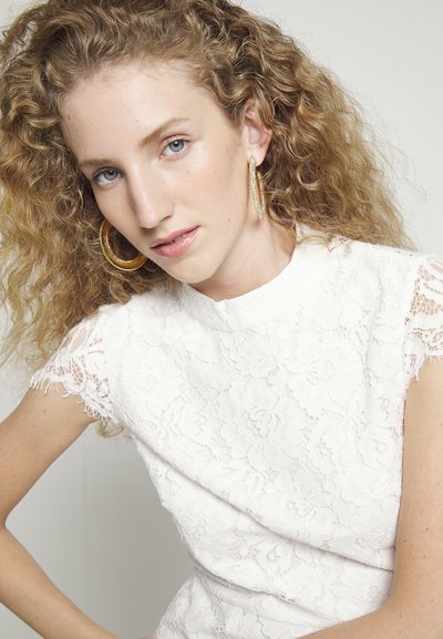 Λευκή δαντελένια μπλούζα με κοντό μανίκι, που διαθέτει ψηλό λαιμό και λουλουδάτο σχέδιο. Μεγάλες χρυσές σκουλαρίκια σε σχήμα κρίκου τονίζουν την εμφάνιση. Ορατά σγουρά μαλλιά.