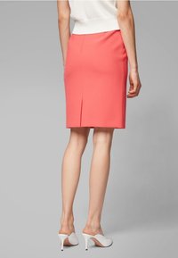 Jupe crayon corail avec une fente à l'arrière, ajustée, fabriquée en tissu lisse. Portée avec des talons ouverts blancs.