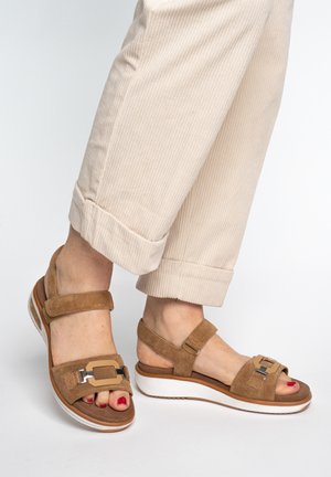Feet wearing beige corduroy pants and tan suede platform sandals with metal buckle details, standing on a white background.