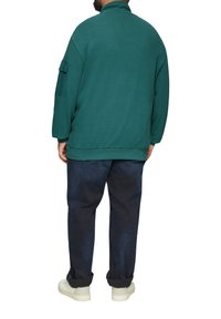 Un sweat-shirt turquoise avec un col montant, des manches longues et une poche à rabat, associé à un pantalon bleu foncé et des baskets de couleur claire.