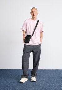 Chemise rose clair à manches courtes, pantalon large gris, sac bandoulière noir et baskets beiges sur un sol en moquette bleue.