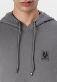 Sweatshirt cinzento com capuz, cordões e um pequeno emblema preto com asas. O material é suave e tem uma textura lisa.
