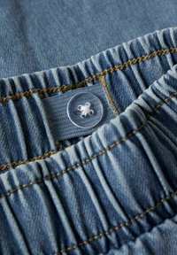 Denim tkanina plave boje, s naboranim elastičnim dijelom i prozirnim plastičnim gumbom s cvjetnim uzorkom. Vidljivo šivanje u zlatnoj boji.