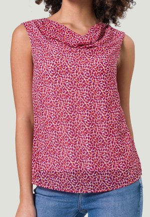 Ærmeløs lyserød bluse med rødt og orange løvmønster, draperet halsudskæring, båret med blå jeans på en person med krøllet hår.