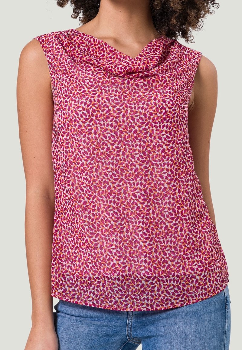 Blouse rose sans manches avec motif de feuilles rouges et orange, col drapé, portée avec un jean bleu sur une personne aux cheveux bouclés.