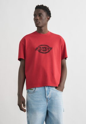 Άντρας που φοράει κόκκινο T-shirt της Dickies και ανοιχτόχρωμα τζιν, στέκεται με το ένα χέρι στην τσέπη μπροστά σε απλό φόντο.