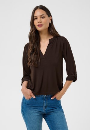 Jeune femme aux longs cheveux foncés porte un chemisier marron foncé à col en V avec des manches retroussées et un jean bleu, debout les mains dans les poches.