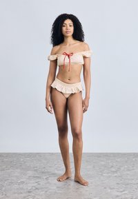 Μπικίνι με τους ώμους ακάλυπτους σε ανοιχτό καφέ χρώμα με κόκκινες πουά, με βολάν στα άκρα και κόκκινη λεπτομέρεια δέσιμο μπροστά.