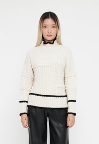 Krämfärgad kabelstickad tröja med svarta accenter, hög krage och texturerade mönster. Har en sidoficka och ribbade ärmslut och nederkant.