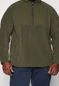 Uomo che indossa un pullover in pile verde oliva con una cerniera a metà e pantaloni blu navy, mostrando solo il torso e le braccia.