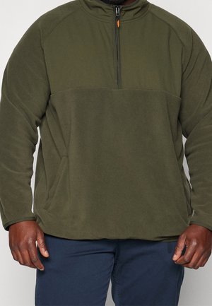 Man wearing olive green fleece pullover with a half zipper and navy blue pants, showing torso and arms only.