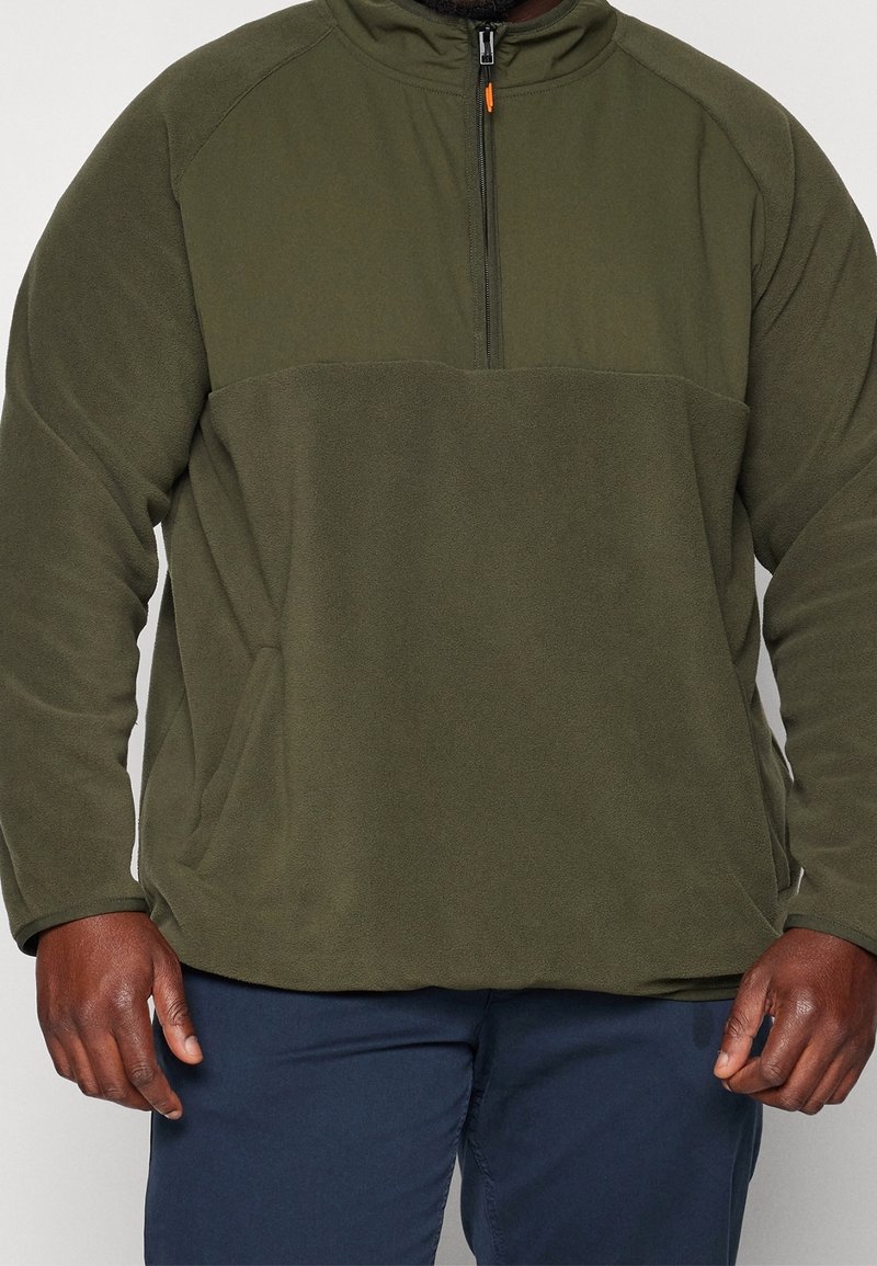 Uomo che indossa un pullover in pile verde oliva con una cerniera a metà e pantaloni blu navy, mostrando solo il torso e le braccia.