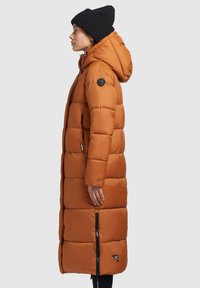 Lang, puffy jakke i orange med hætte. Har sorte knapdetaljer og en sidelukning. Bæres med en sort strikket beanie.