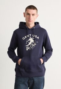 Sudadera azul marino de algodón con un bolsillo frontal, que presenta una impresión gráfica blanca de "GANT USA Ski Club" y acentos de diseño relacionados con el esquí.